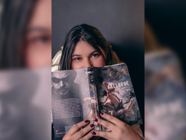 ZoeyGrey with black hair, hiding behind a book with a cover featuring a man's face and the word "Melalaria". She's looking at the camera with dark red nails.