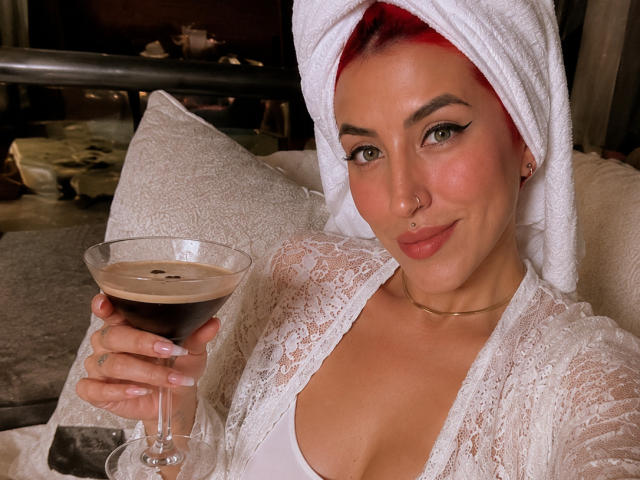 Alissa Brown, met rood haar, een witte handdoek op haar hoofd, een kantjapon, die een glas zwarte koffie vasthoudt, met een lichte glimlach, een neusring en lichte make-up, zittend op een bank..