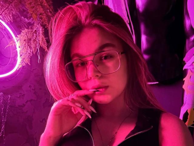 VanesaLucic, con el cabello con toques rosados, usando gafas y una chaqueta negra con cremallera, mordiéndose el dedo con una mirada seductora, iluminación rosa neón y uñas largas..