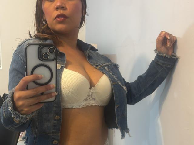SophiaQueens, con piel morena, cabello castaño y labial, vistiendo una chaqueta vaquera sobre un sujetador de encaje blanco, tomando una selfie frente al espejo, mostrando sus pechos, sosteniendo un teléfono negro..