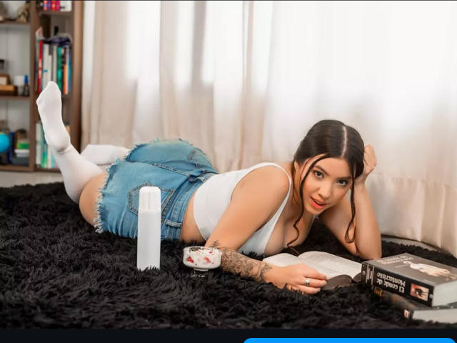 SophiiaRios acostada sobre una alfombra negra de pelo, con una camiseta blanca, pantalones cortos vaqueros azules, calcetines blancos, tatuajes en el brazo derecho, cabello negro, un libro, un pincel y una taza frente a ella..