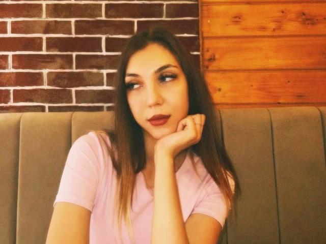 FarahSings mit langen, braunen Haaren, einem pinken Hemd und vollen Lippen, lehnt ihr Kinn in die Hand und blickt zur Seite, in einer grauen Kabine. Ihr Hintern ist nicht sichtbar..