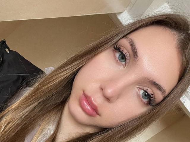 FarahSings tiene el pelo largo y castaño, ojos verdes y labios rosados y brillantes. Lleva puesto un conjunto negro con una prenda blanca debajo, y mira directamente a la cámara con una sonrisa sutil y sensual..