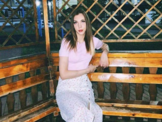 FarahSings com cabelos castanhos longos, camisa rosa e saia branca com estampas florais, apoiada em um banco de madeira, com os braços descansando nas costas, olhando para a câmera, noite lá fora, fundo com treliça de madeira..