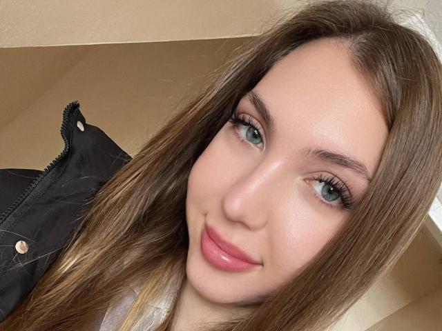 FarahSings heeft bleke huid, groene ogen, lang bruin haar, roze lippenstift en een lichte glimlach. Ze draagt een zwarte jas met een rits..