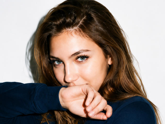 FarahSings, avec ses yeux bleus, ses cheveux bruns et une expression sérieuse, mordille sa main. Elle porte un pull bleu foncé, et sa main recouvre légèrement sa bouche..