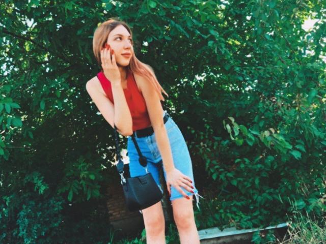 FarahSings met lang bruin haar, een rode top, blauwe shorts met hoge taille, een zwarte tas, staand, hand in de buurt van de mond, groene, bladerrijke achtergrond, kijkt opzij, glimlacht licht, zomerse sfeer..