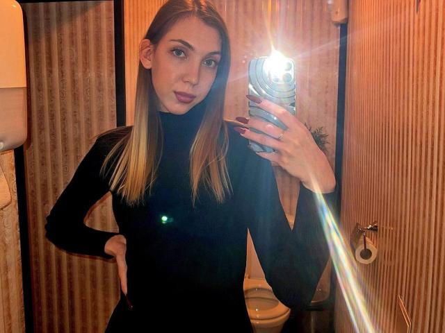 FarahSings, avec ses cheveux blonds, portant un pull noir à col roulé, prend un selfie devant un miroir avec le flash, montrant une silhouette mince et des fesses petites et discrètes, dans une salle de bain..