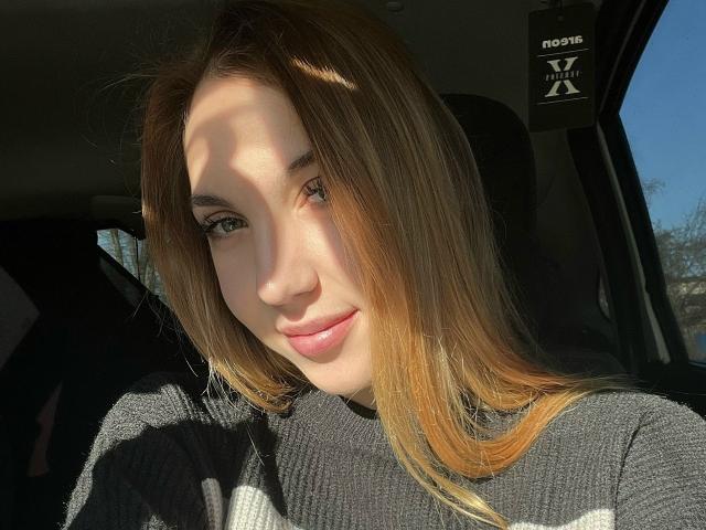 FarahSings tiene el cabello castaño claro, ojos verdes y una cara linda con una ligera sonrisa, vestida con un suéter blanco y negro, iluminada por el sol dentro de un coche..