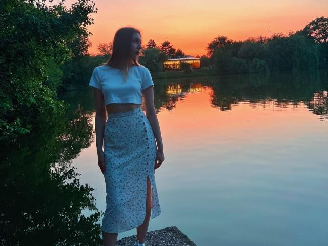 FarahSings se tient au bord d'un lac au coucher du soleil, vêtue d'un crop top blanc et d'une jupe fleurie fendue sur la cuisse, et regarde sur le côté. Ses fesses sont légèrement visibles..