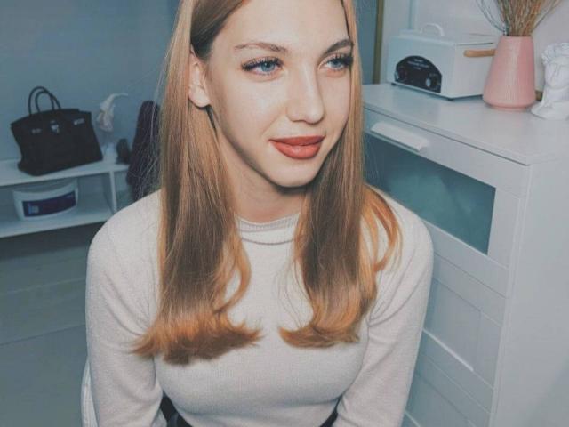 FarahSings, con i capelli biondi, gli occhi azzurri e il rossetto rosso, indossa un maglione bianco, ha un sorriso carino e ciglia seducenti, ed è seduta in una stanza con un vaso rosa..