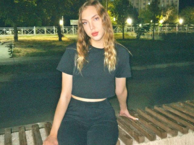 FarahSings met golvend blond haar, rode lippenstift, een zwart topje en zwarte broek, zittend op een houten bank in de nacht, met haar hand op de zitting, en die lichtelijk opzij kijkend..