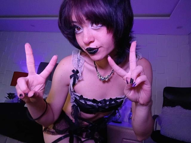 ViolettClay with short black hair, white face paint, black lipstick, and lace bra, making double peace signs, showing small tits, and wearing a black necklace in a purple-lit room.
