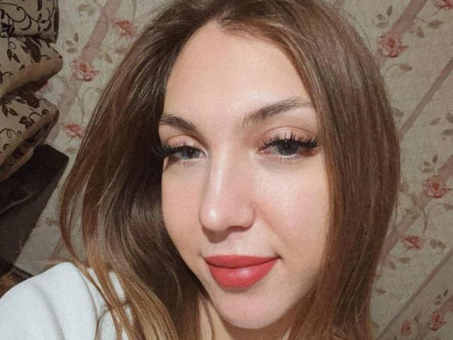 FarahSings heeft lang bruin haar, blauwe ogen en rode lippenstift. Ze glimlacht met lange wimpers, draagt een witte top en heeft een schattig, mooi gezicht..