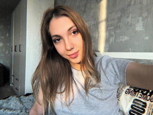 FarahSings, com seus longos cabelos castanhos, camiseta cinza clara e um sorriso discreto, exibe seios pequenos e um rosto adorável, em um quarto iluminado pelo sol. Ela está segurando uma câmera para tirar uma selfie..
