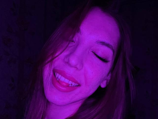 FarahSings a les yeux fermés, souriant avec ses lèvres rouges et ses dents visibles, sous un éclairage violet, avec ses cheveux bruns qui couvrent partiellement son visage..