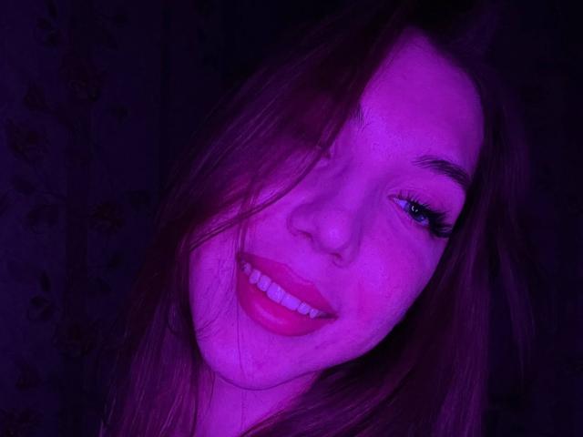 FarahSings a un visage éclairé de violet, de longs cheveux bruns, des yeux bleus et du rouge à lèvres, et elle sourit en montrant ses dents blanches. Ses fesses ne sont pas visibles, mais son visage est bien en évidence..