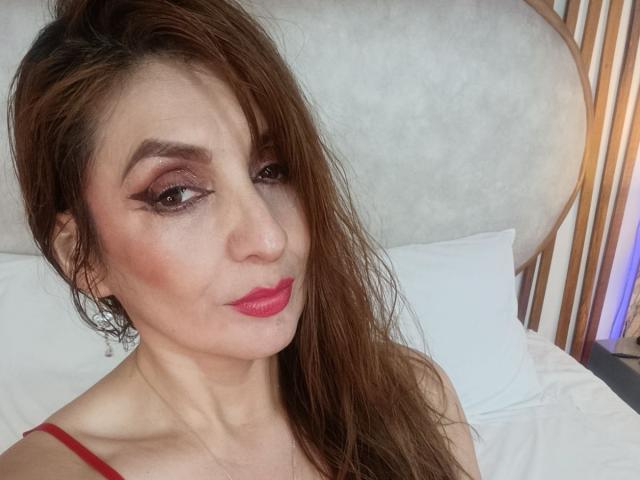 Elena Hart tem cabelos castanhos e úmidos, batom rosado-avermelhado e maquiagem esfumada nos olhos. Ela está olhando para a câmera com um leve sorriso, vestindo uma blusa vermelha que mostra seus seios..