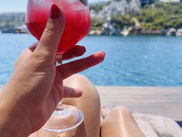 La mano di BadAngel, con le unghie dipinte di rosso, tiene un bicchiere da cocktail rosso, con uno sfondo di acqua blu illuminata dal sole. Le gambe abbronzate e lucide di BadAngel sono in primo piano..