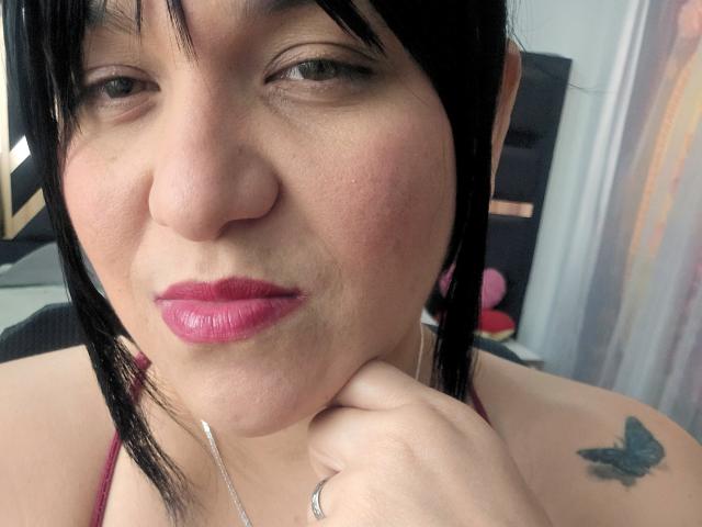 VivianaThomson has pink lipstick, black hair, and a butterfly tattoo on her shoulder. She's resting her chin on her hand with a slight pout. Her nose is small.