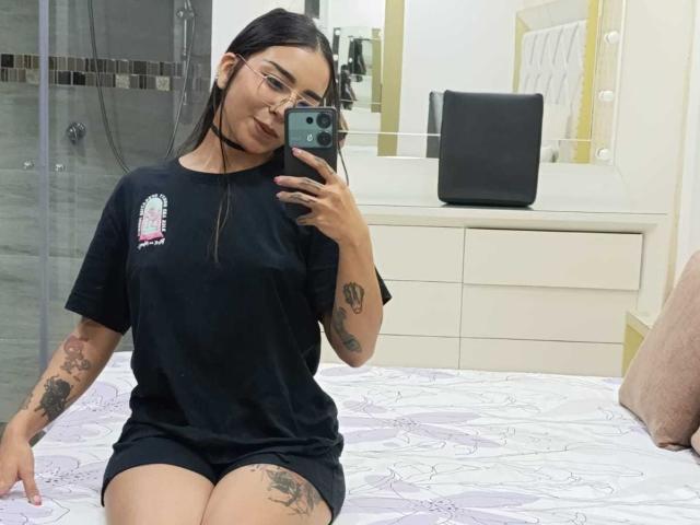 MilaThorne, com cabelos pretos longos, óculos e tatuagens, vestindo uma camiseta preta e shorts, sentada em uma cama, tirando uma selfie no espelho com um telefone verde..