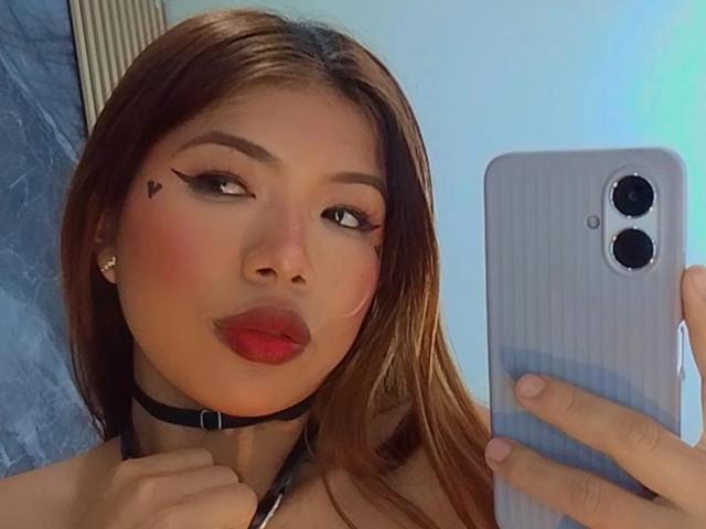 CloeLombardx with red lips, black choker, and heart tattoo on cheek, holding a phone, brown hair, and looking at the camera, wearing small earrings.