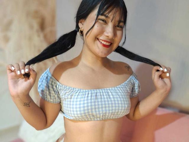 AlejaMiller with pigtails, smiling, wearing a blue and white checkered crop top, showing small tits, tattoos on arm and chest, holding pigtails, red lipstick, ear gauges.