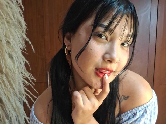 AlejaMiller with black pigtails, red lipstick, and small tattoo on right shoulder, biting finger, looking at camera, wearing off-shoulder top, gold hoop earrings, wooden background.