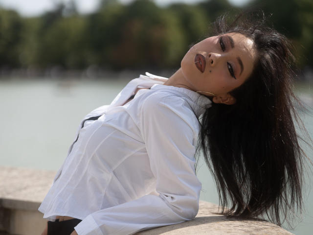 Nashiro Sakurai, con lunghi capelli neri, appoggiata a un muro, indossa una camicia bianca con bottoni, occhi chiusi, testa inclinata verso l'alto, rossetto scuro, piccolo tatuaggio sul viso, acqua sullo sfondo..