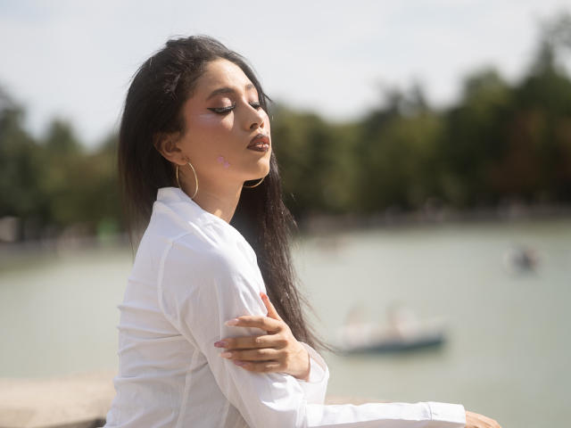 Nashiro Sakurai, met lang donker haar, draagt een witte blouse, armen gekruist, ogen gesloten, gouden oorringen, staat bij een meer, zacht zonlicht, kalme uitdrukking, handen op de armen..