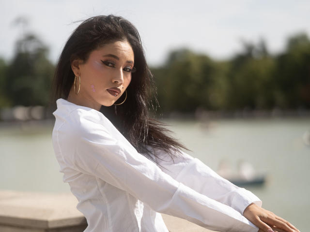 Nashiro Sakurai, met lang zwart haar, gekleed in een witte blouse, met gouden oorringen en roze lippenstift, staat bij een waterweg en kijkt over haar schouder met een serieuze uitdrukking..
