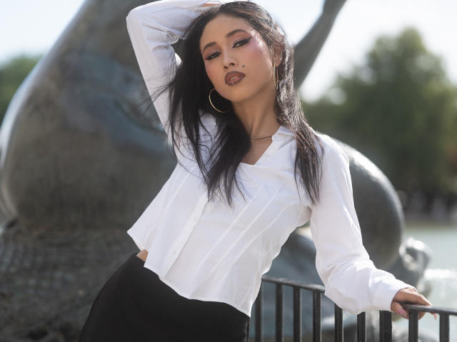 Nashiro Sakurai, met lang zwart haar, een witte blouse, een zwarte rok en oorringen, leunt tegen een metalen hekwerk, met haar hand in haar haar, en kijkt verleidelijk naar de camera..