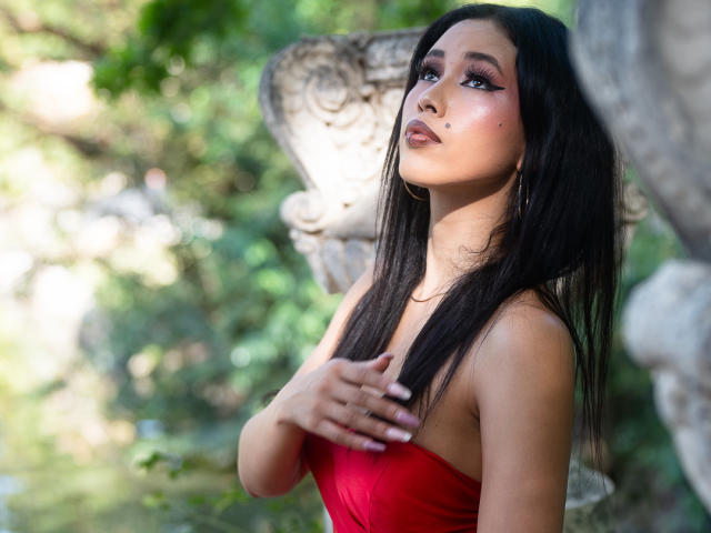 Nashiro Sakurai met lang zwart haar, een rode jurk zonder bandjes, en roze eyeliner, kijkt omhoog, met haar hand op haar borst, waarbij haar borsten licht zichtbaar zijn, staand in de buurt van een stenen sculptuur, en ze ziet er aantrekkelijk uit..