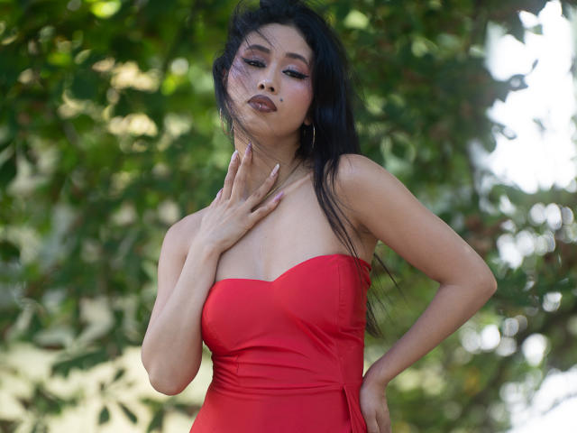Nashiro Sakurai met lang zwart haar, een rode jurk zonder bandjes, haar hand op haar borst, volle lippen, paarse oogschaduw en een zelfverzekerde pose, waarbij haar borsten en schattige gezicht te zien zijn..