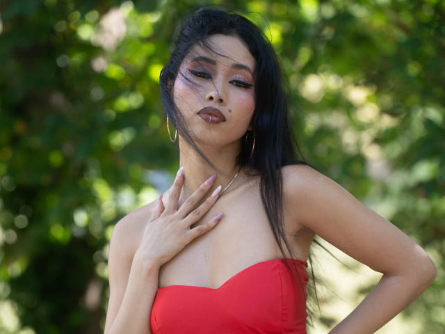 Nashiro Sakurai, met lang zwart haar, draagt een rode top zonder bandjes, heeft haar hand op haar borst, toont haar borsten en kijkt omhoog met roze eyeliner en oorringen..