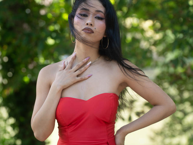 Nashiro Sakurai, met lang zwart haar, draagt een rode jurk zonder bandjes, gouden oorringen en paarse nagellak, en raakt haar borst aan. Ze kijkt zelfverzekerd, met een lichte bries in haar haar..