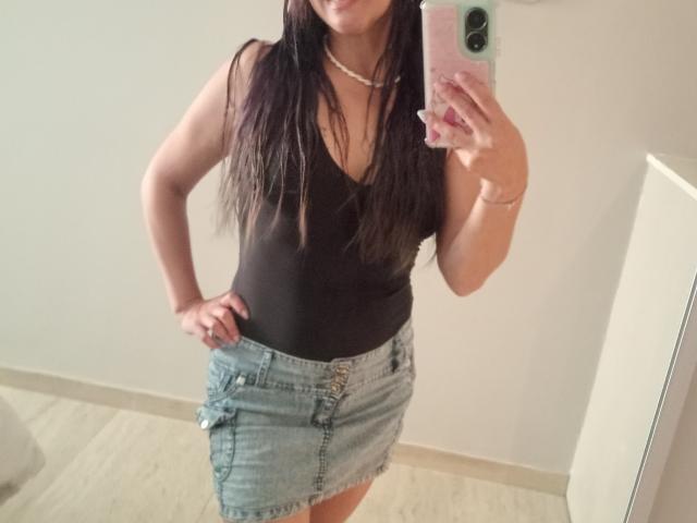 Crisht, avec de longs cheveux bruns, un débardeur noir et une jupe en jean bleu, se prend en photo devant un miroir, une main sur la hanche, portant un collier, avec un téléphone dans une coque rose, sans seins visibles..