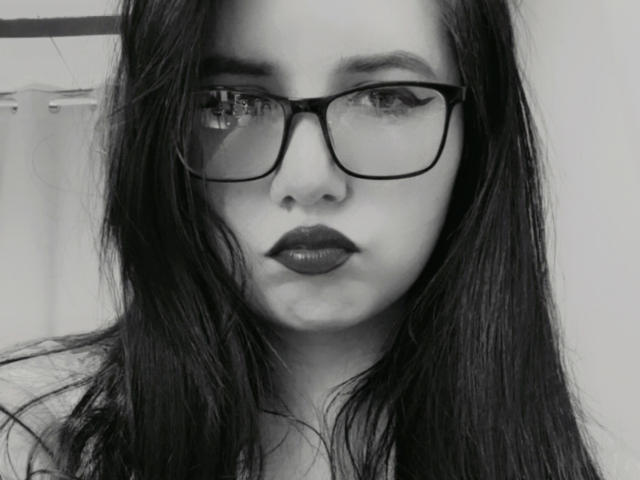 Fotografia in bianco e nero di Kkatalina con lunghi capelli scuri, che indossa occhiali spessi, rossetto scuro e un'espressione seria, che mostra il suo viso e la parte superiore del petto. Il suo seno è parzialmente visibile..