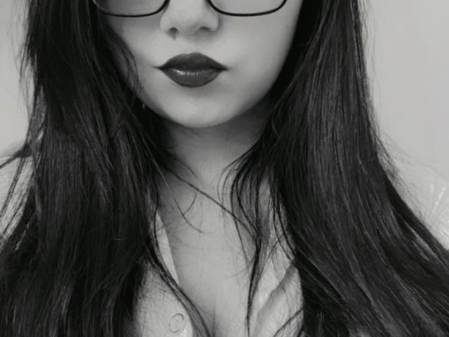 Kkatalina's face with dark lipstick, long black hair, glasses, and a partially open shirt showing her tits. Black and white image.