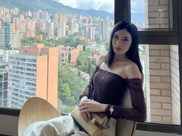 Fiorella Leroy, con cabello negro largo, una blusa marrón con un hombro descubierto y pantalones blancos, sentada junto a una ventana, sosteniendo un bolso de paja, con un paisaje urbano en el exterior y montañas al fondo..
