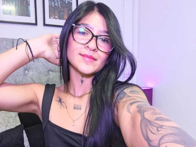 LuciadelaVega with long black hair, glasses, and pink lipstick, showing off tattoos on her arm and chest, wearing a black tank top, smiling with a hand in her hair.