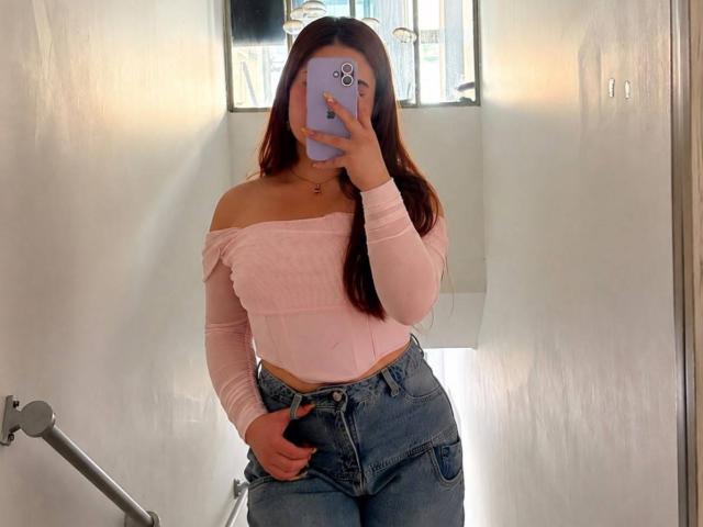 VicluX, com cabelos castanhos longos, uma blusa rosa com um ombro de fora e jeans azuis, segurando um telefone roxo que cobre seu rosto, está em um corredor claro com paredes brancas..