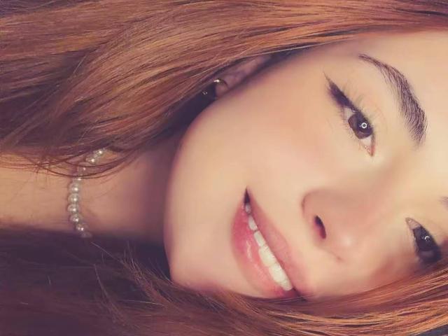 O rosto de VicluX, com cabelo castanho, nariz pequeno e dentes brancos, usando um colar de pérolas, tem um leve sorriso, e uma piercing preta na orelha direita..
