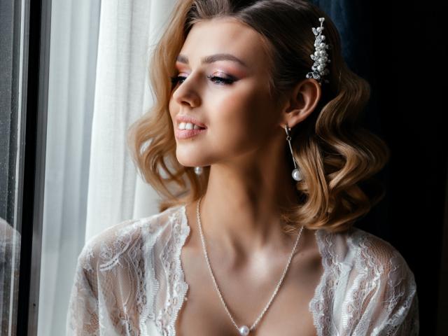 Margorie Symon, com cabelos loiros ondulados, vestindo um vestido de renda, brincos de pérolas e um acessório floral no cabelo, sorrindo suavemente, olhando para fora de uma janela. Seios visíveis sob a renda..