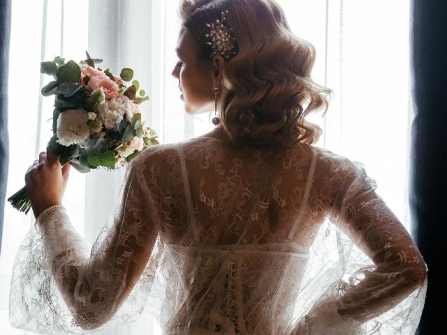 MargorieSymon, com cabelos loiros ondulados, vestido de noiva de renda, segurando um buquê rosa e branco, de costas para a câmera, luz branca atrás, acessório de pérolas no cabelo, elegante, suave, delicada..