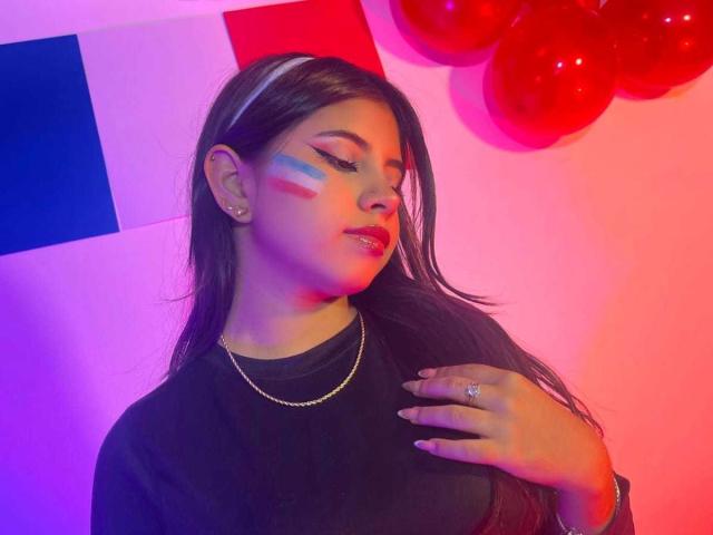 Celeste Morel met geschminkt gezicht, rode en blauwe strepen, draagt een zwarte top, een gouden ketting, een ring, heeft lang bruin haar, er is roze en paarse verlichting, rode ballonnen, haar ogen zijn gesloten, haar hand rust op haar borst..