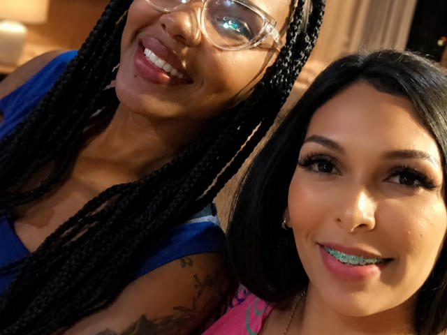 Brenda mit Zöpfen und Brille, lächelnd mit herausgesteckter Zunge, steht neben Natalya mit schwarzem Haar, Zahnspange und einem pinken Oberteil. Beide haben Tattoos und machen ein Selfie..