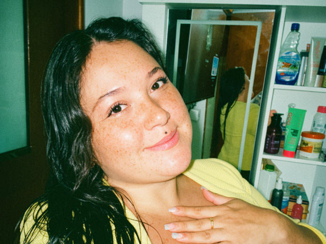 ColetteBessette tem sardas, um piercing no nariz e cabelo escuro ondulado, vestindo uma camisa amarela, sorrindo com a mão no peito, em uma selfie no espelho..