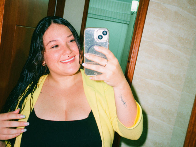 Colette Bessette, con el cabello negro y mojado, sonriendo, sosteniendo un teléfono, con un cárdigan amarillo, una blusa negra, mostrando un busto grande, un tatuaje pequeño en el brazo derecho, tomando una selfie en el espejo, uñas verdes..