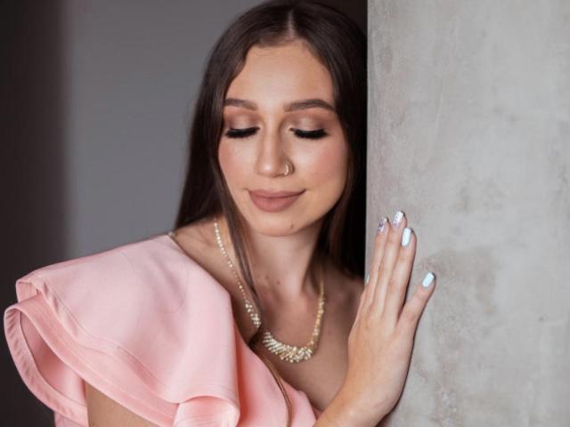 Maittee, avec ses longs cheveux bruns, sa robe rose et son collier de perles, appuyée contre un mur gris, les yeux fermés, souriante, montrant ses ongles peints en bleu, et portant un anneau au nez..